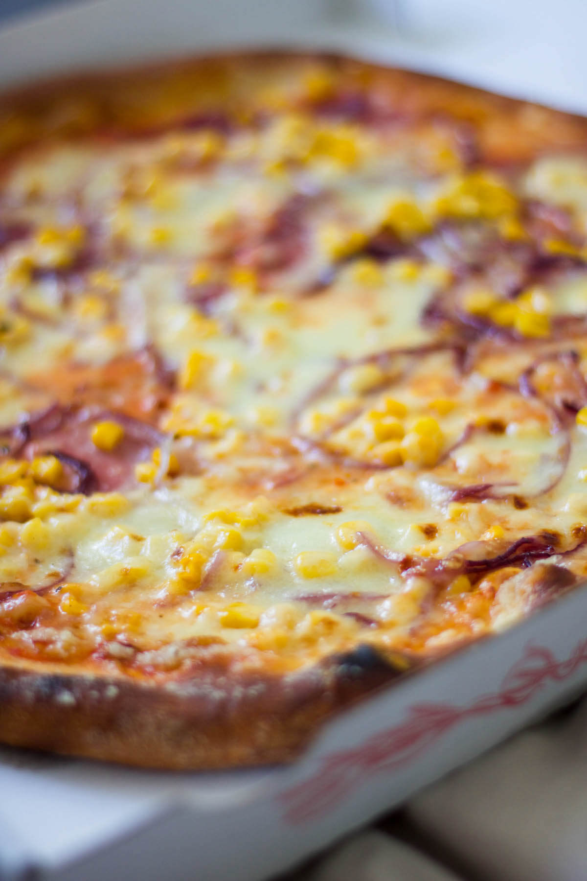 Pizza mit Schinken, Käse, Mais und Zwiebel in Pizzakarton am Bett